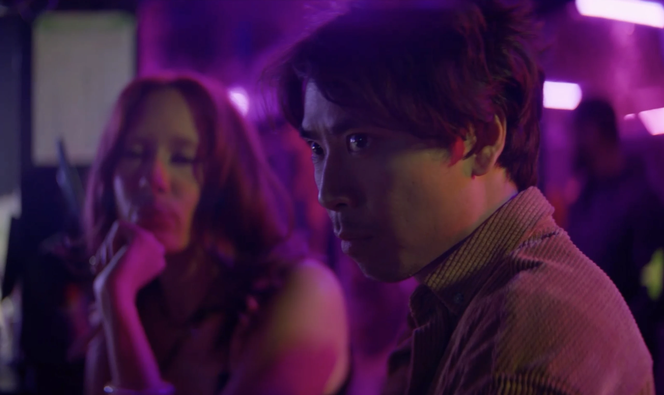 A man turns toward the camera beside a woman in a magenta-lit interior.
