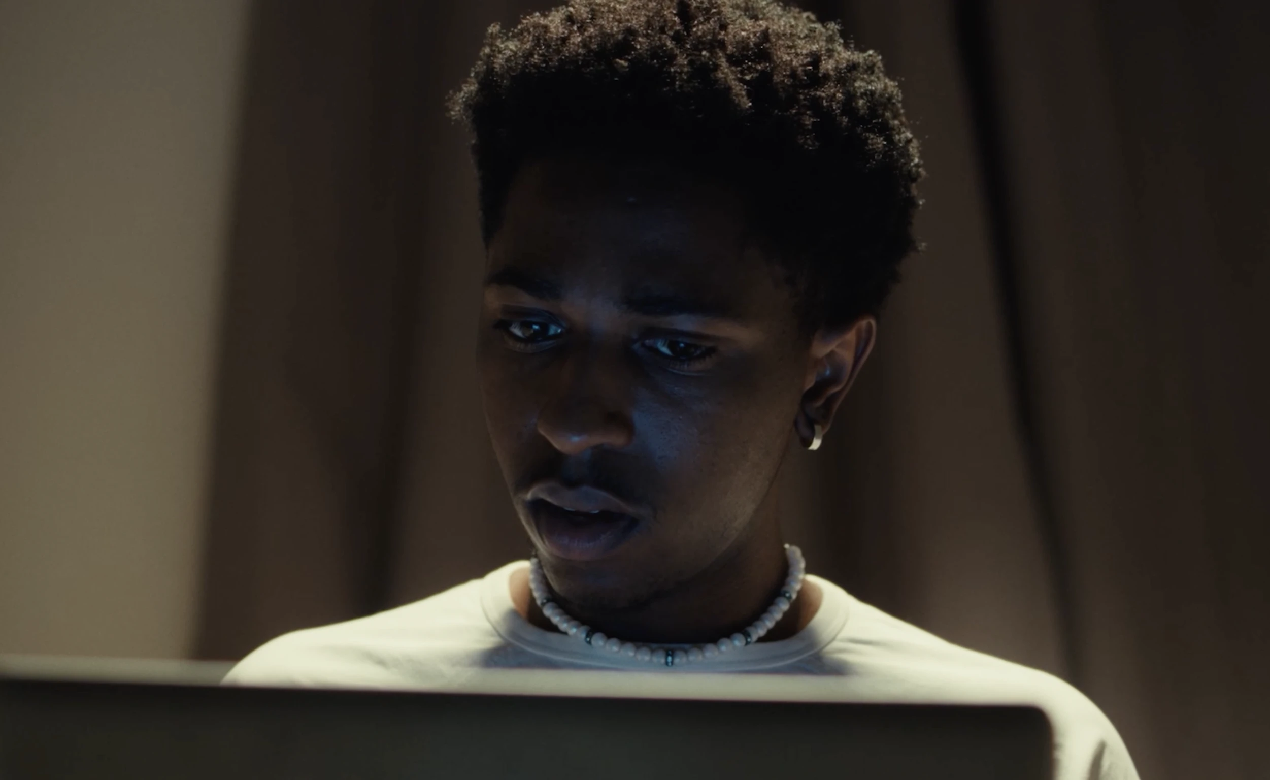 A man wearing a beaded necklace looks down at a laptop in a dimly lit room.